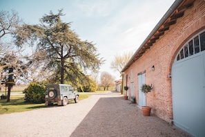 A gravel driveway leading to a brick building with a garage door, a parked vehicle, and trees in the background.
