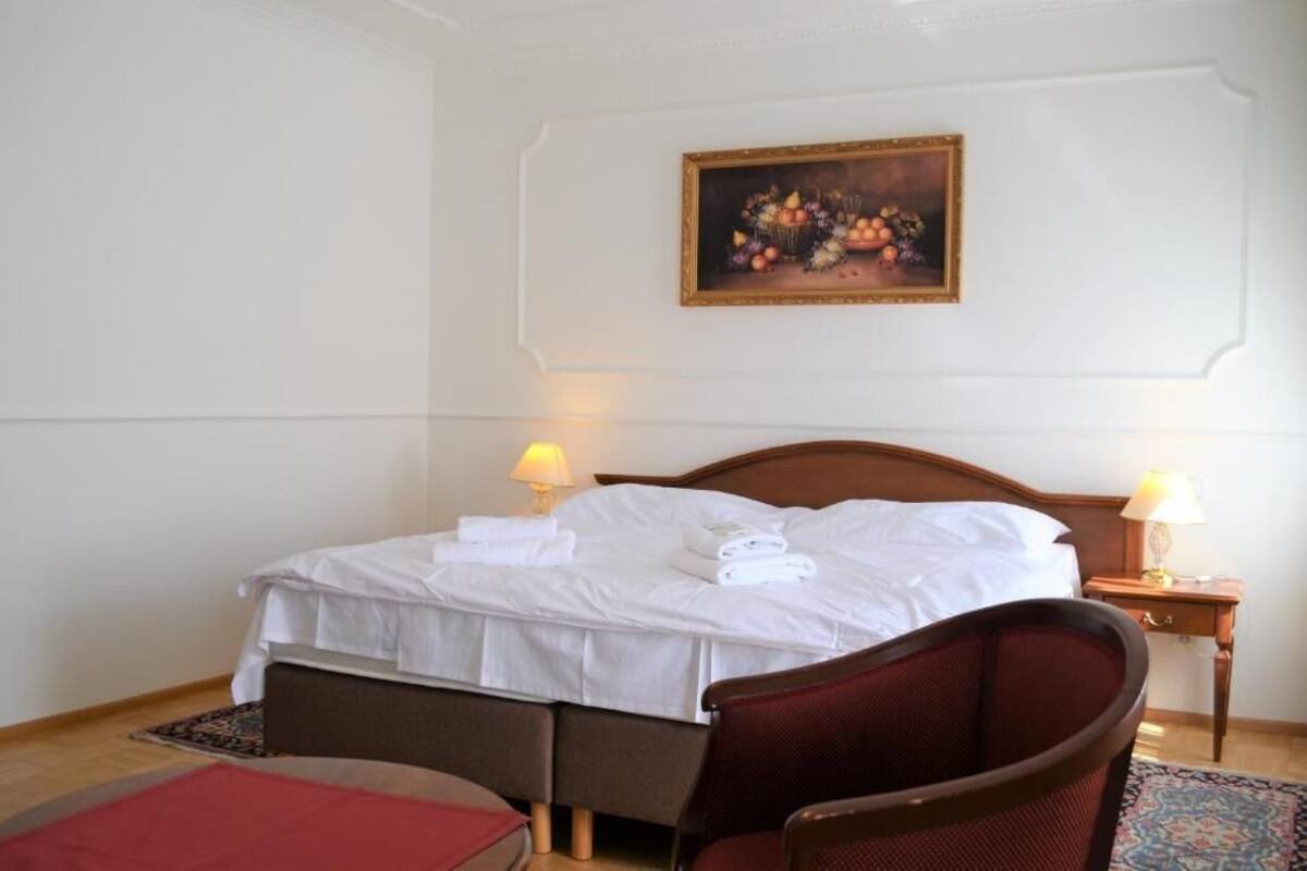 A neatly made bed with white linens and towels, a wooden headboard, a red armchair, a small wooden nightstand with a lamp, and a framed painting of fruit on the wall.