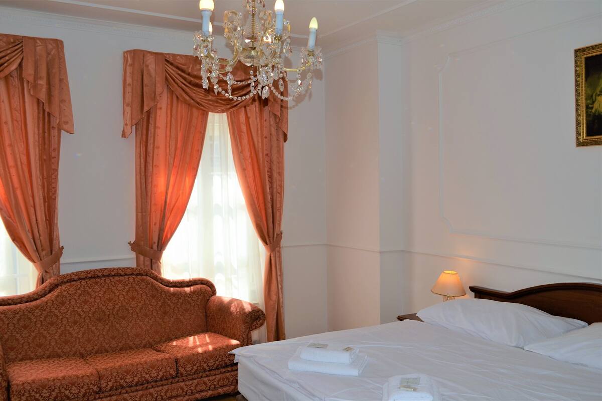 A hotel room with a bed, a sofa, a chandelier, and curtains.