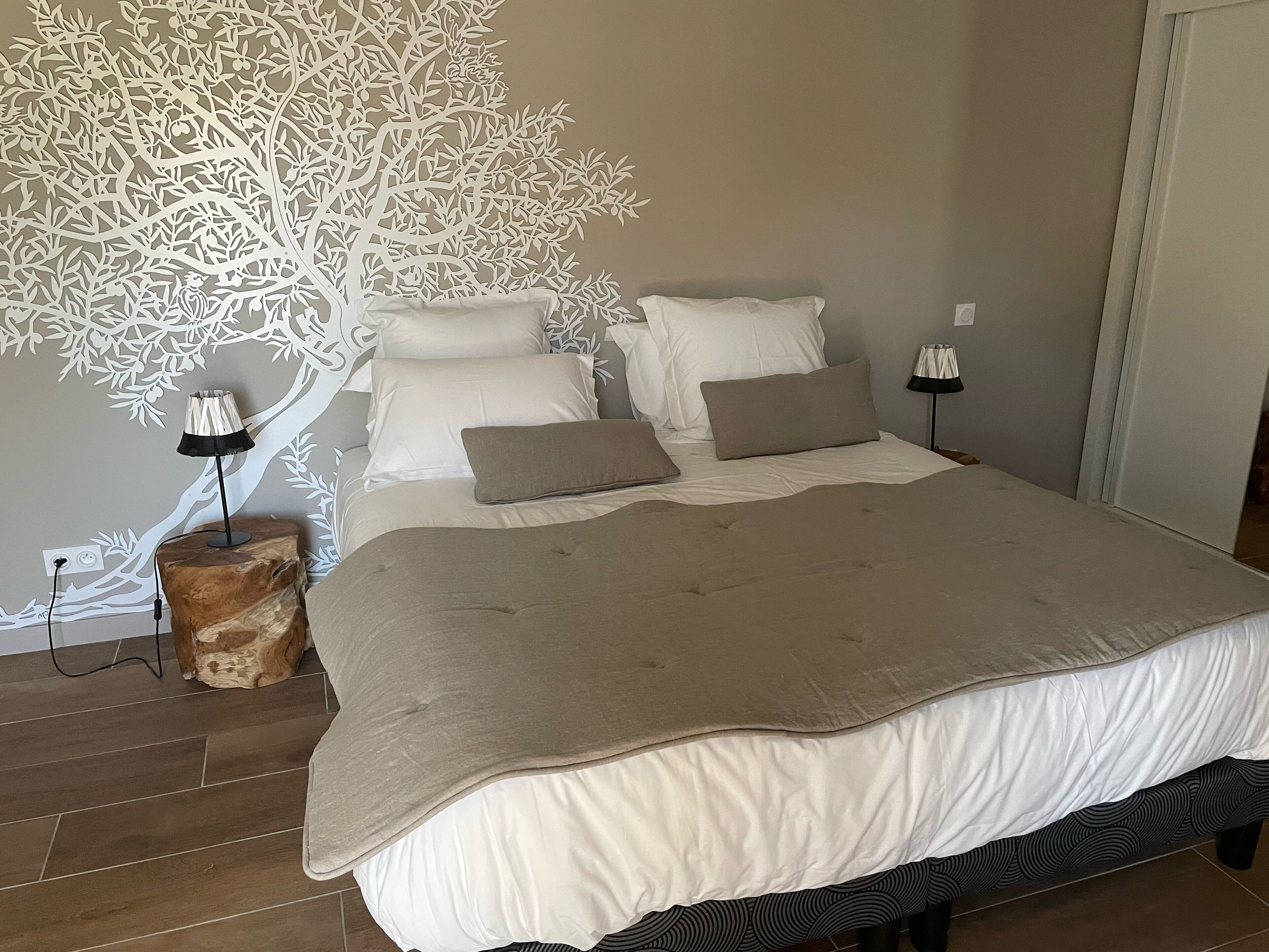Une chambre avec un grand lit, une fresque représentant un arbre sur le mur, deux lampes de chevet et une table de nuit en bois.