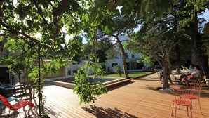 Una terraza de madera al aire libre con zonas de estar, rodeada de árboles y un edificio al fondo.