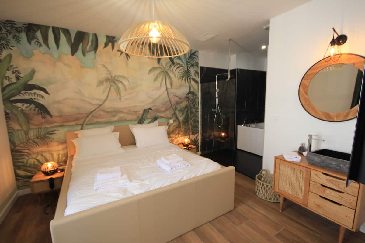 A bedroom with a large bed, a nightstand with a lamp, a wooden dresser, a mirror, and a tropical mural on the wall.