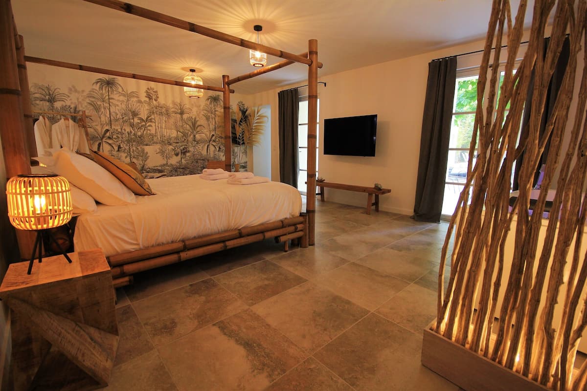 A bedroom with a large bed, a bedside lamp, a television, and a view of the outdoors.