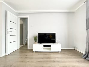 Una sala de estar moderna con un televisor de pantalla plana sobre un mueble blanco, suelo de madera y una puerta que conduce a otra habitación.