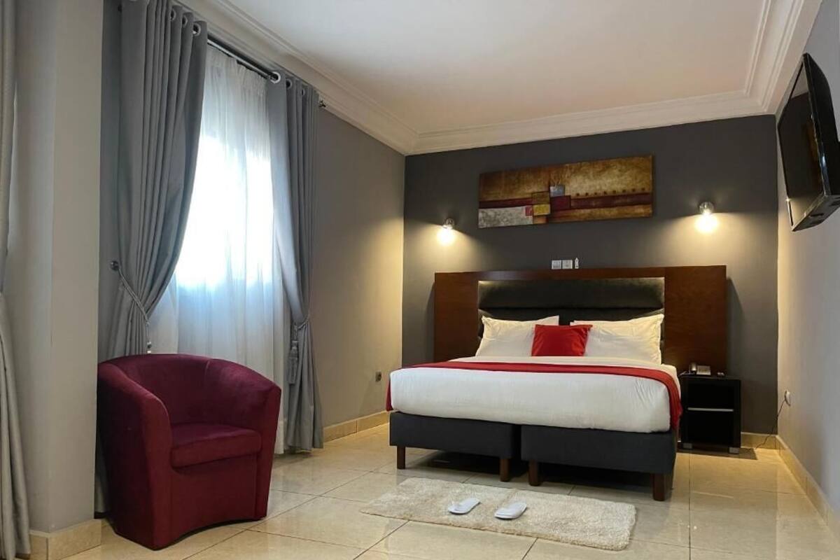 A hotel room with a bed, a red armchair, a TV, and a painting on the wall.
