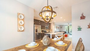 A modern kitchen with a wooden dining table set for four, a hanging light fixture, and a wall-mounted sign spelling "EAT". - 3BR Resort Pool + Jacuzzi Minutes to Disney 4730 (Kissimmee)