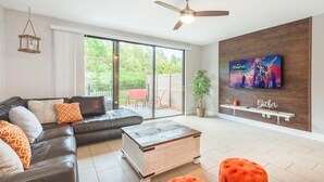A modern living room with a sectional sofa, a coffee table, and ottomans. There is a TV mounted on the wall, a ceiling fan, and a sliding glass door leading to an outdoor area. - 3BR Resort Pool + Jacuzzi Minutes to Disney 4730 (Kissimmee)