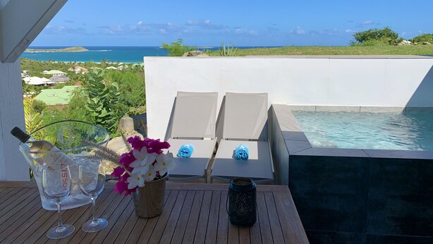 A wooden table with two wine glasses, a pitcher, and a vase with flowers. A lantern is on the table. A pool with a white wall and a view of the ocean and greenery is in the background. - SUNSEEKER SXM (Marigot)