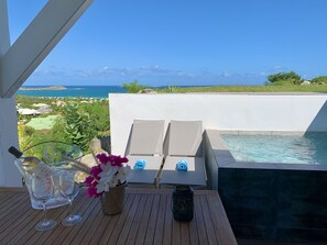 Un tavolo di legno con due bicchieri da vino, una brocca e un vaso con fiori. Sul tavolo c'è anche una lanterna. Sullo sfondo, una piscina con un muro bianco e vista sull'oceano e sulla vegetazione.