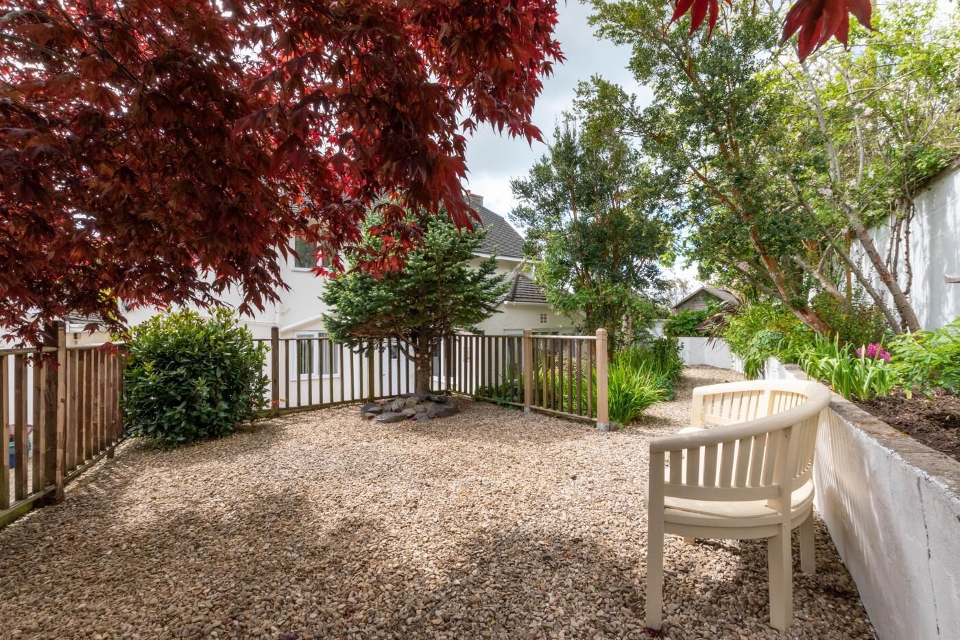 A gravel patio with a wooden fence, a small tree, and a bench.