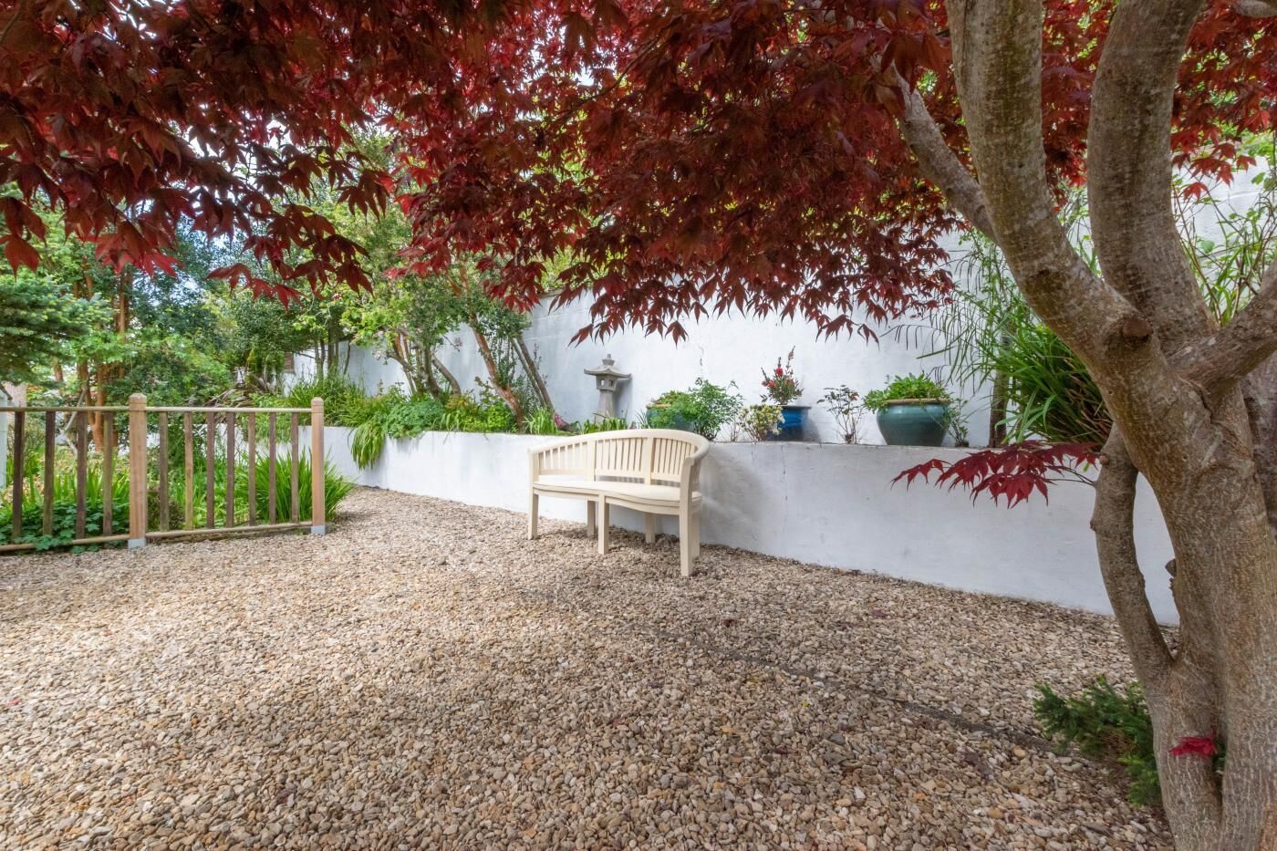Ein Garten mit einer Bank, Kiesstreu und Kübelpflanzen vor einer weißen Wand.