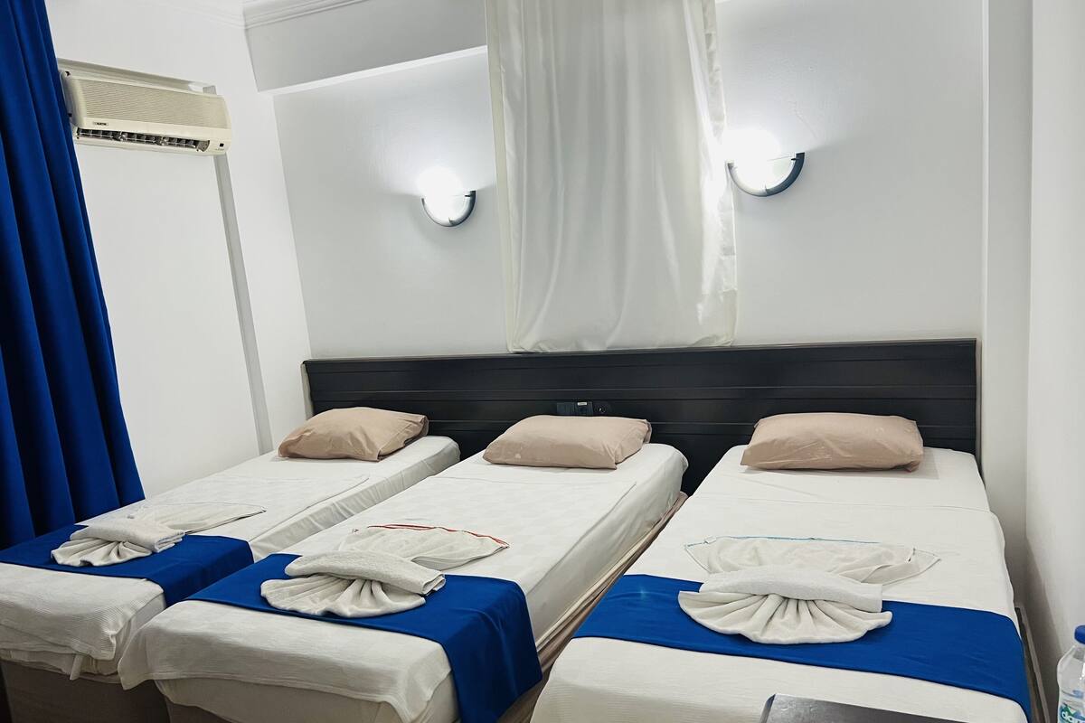 A hotel room with three single beds, each with a blue and white bedspread, and beige pillows. There are two wall-mounted lights and a ceiling fan.