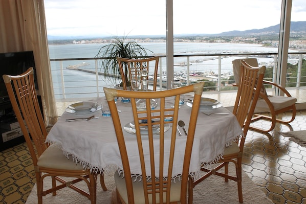 A dining area with a table set for four, overlooking a body of water and a cityscape.