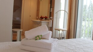Una cama bien hecha con toallas blancas dobladas y una pequeña decoración floral. Se observa una silla blanca y una mesita de noche al fondo.