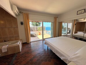A hotel room with a large bed, a bathroom with a bathtub, and a view of the sea through a sliding door.