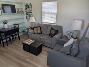 A living room with a grey sofa, a coffee table, a TV on the wall, and a window with blinds.