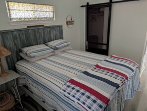 A bedroom with a bed, a bedside table, a lamp, a window with blinds, a wall clock, and a door with a sliding mechanism. - The Pelican at Magnolia Beach
 (Port Lavaca)