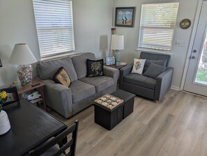 Uma sala de estar com um sofá cinzento, uma mesa de centro e uma luminária. Há duas janelas com persianas, um quadro na parede e um relógio.