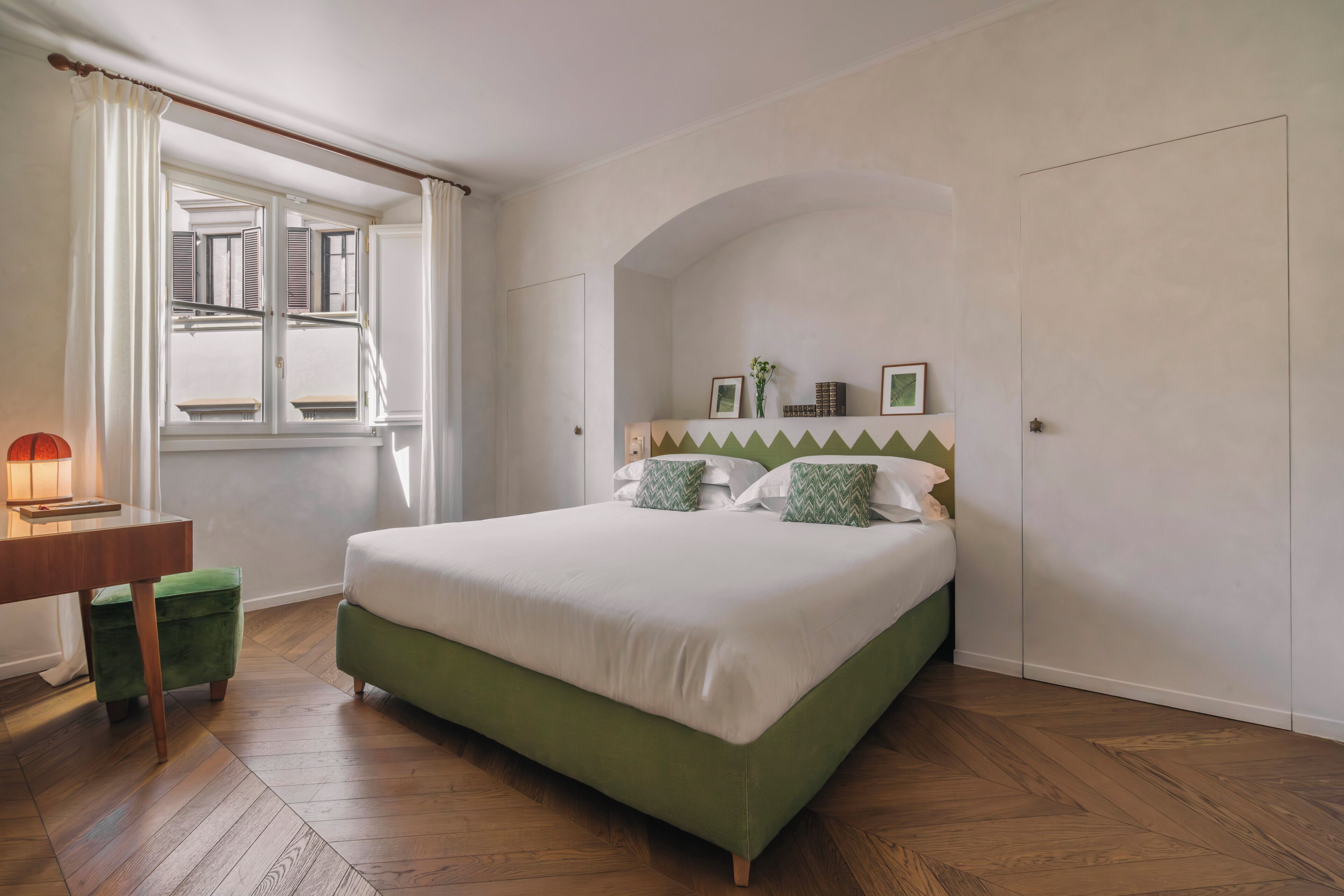 Ein Schlafzimmer mit einem grünen Bett, einem Holztisch, einem grünen Stuhl, einem Fenster mit Vorhängen und einer Wandregal mit Bildern und einer Vase.