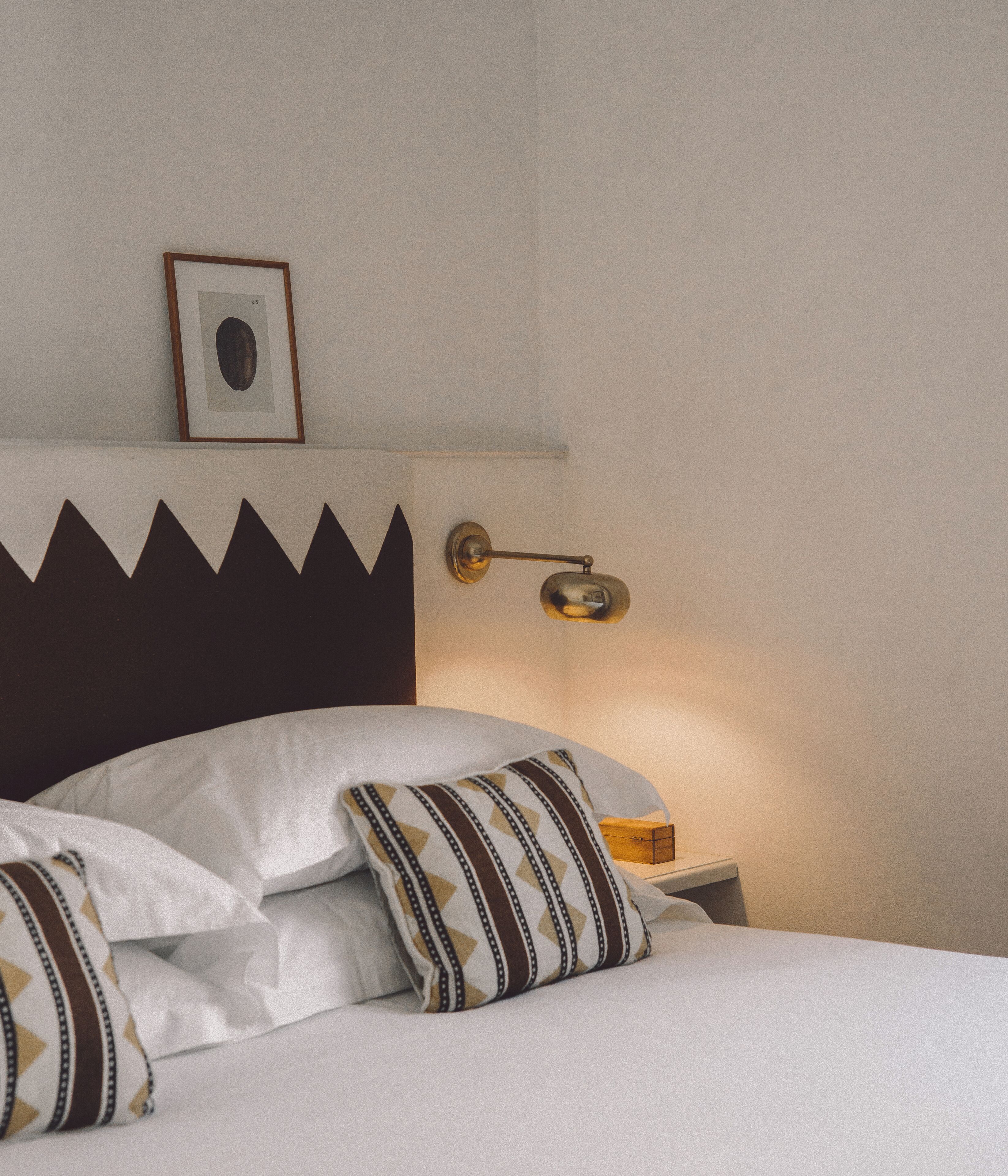 A neatly made bed with white linens and patterned pillows, a framed picture on the wall, and a bedside table with a lamp.