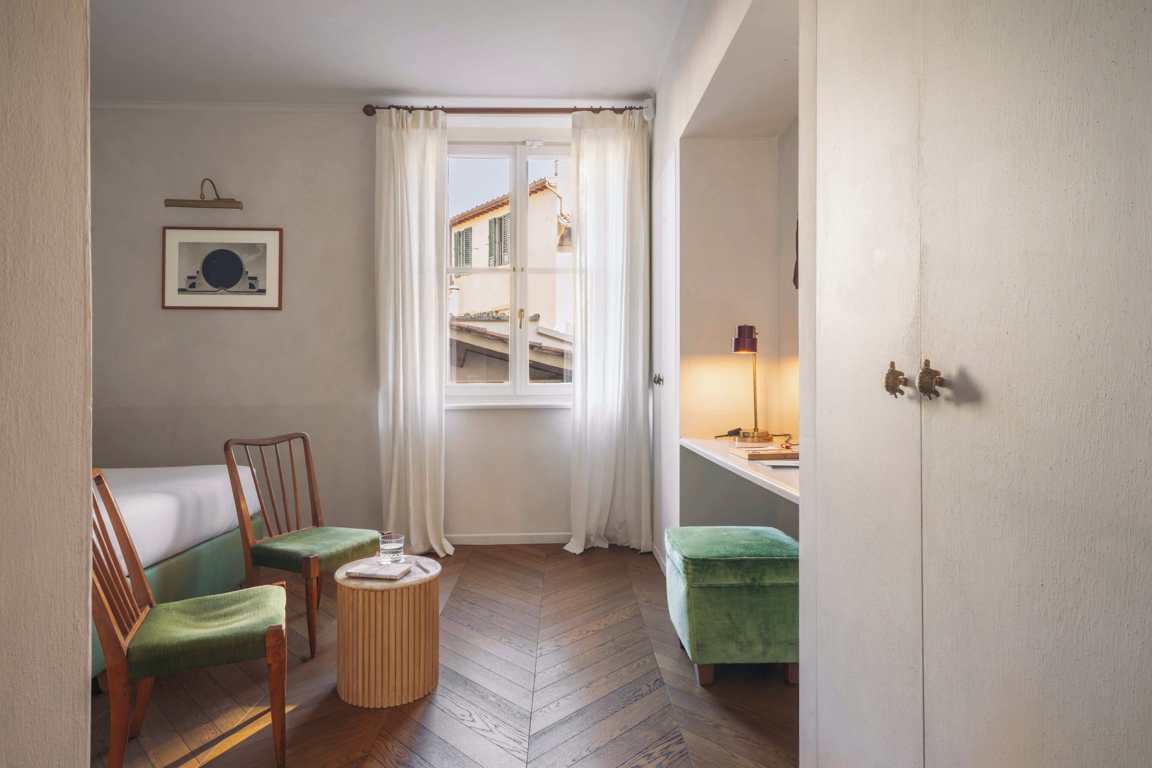 A small, cozy room with a bed, two chairs, a round table, a green ottoman, a lamp, and a window with curtains.