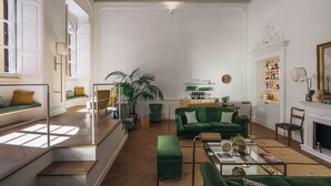Un salon spacieux avec un canapé vert, une table basse et une cheminée. On y trouve une bibliothèque remplie de divers objets et une grande fenêtre qui laisse entrer la lumière naturelle.