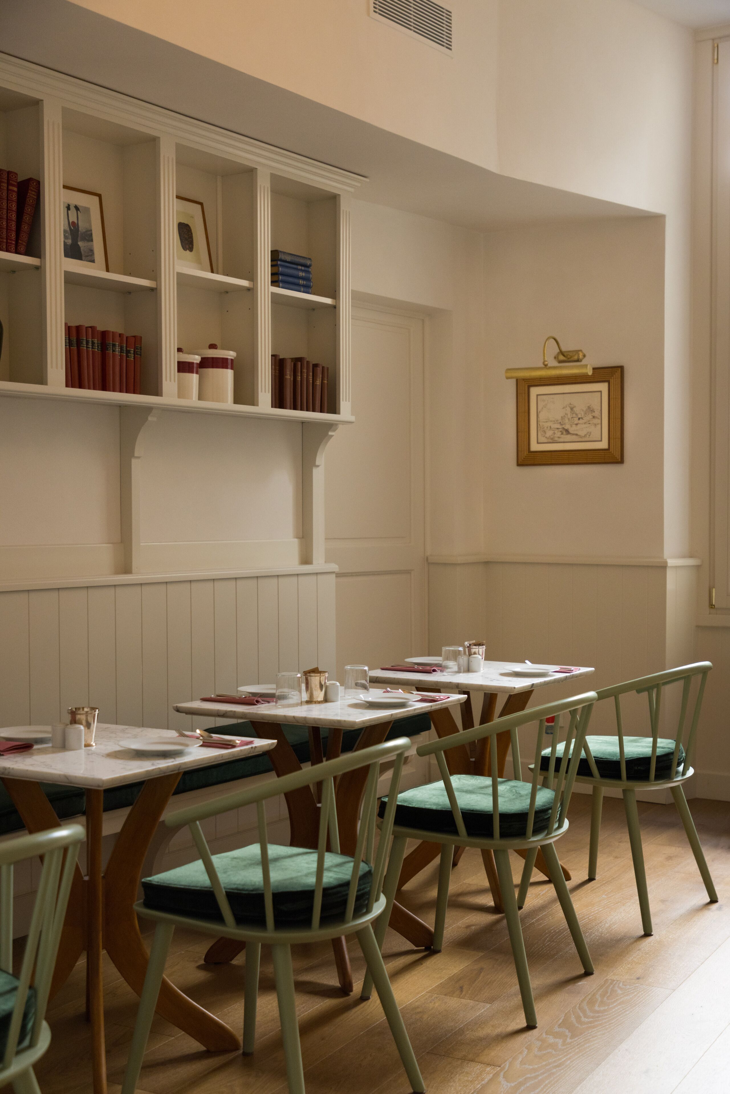 A dining area with wooden tables and chairs, white walls, and shelves with books and framed pictures.