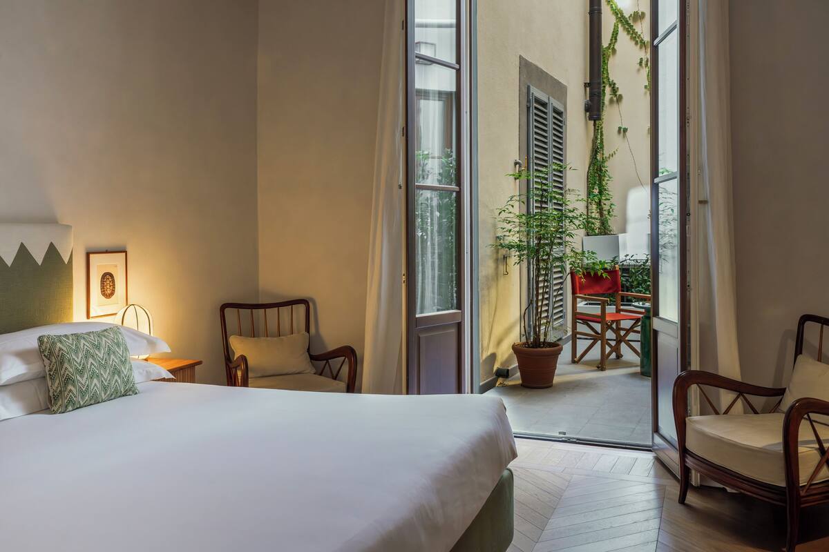 A hotel room with a bed, two chairs, a potted plant, and a view of the outside balcony.