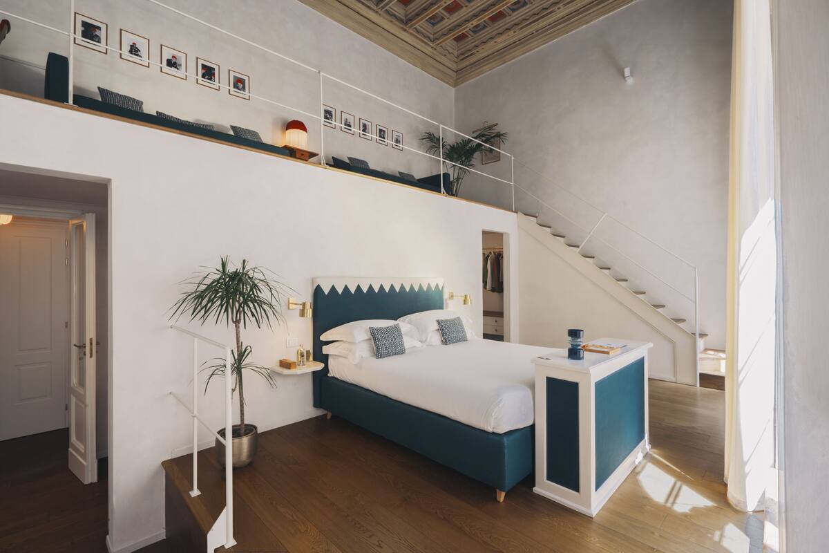 A bedroom with a bed, a bedside table, a nightstand, a potted plant, a staircase, and a ceiling with a decorative pattern.