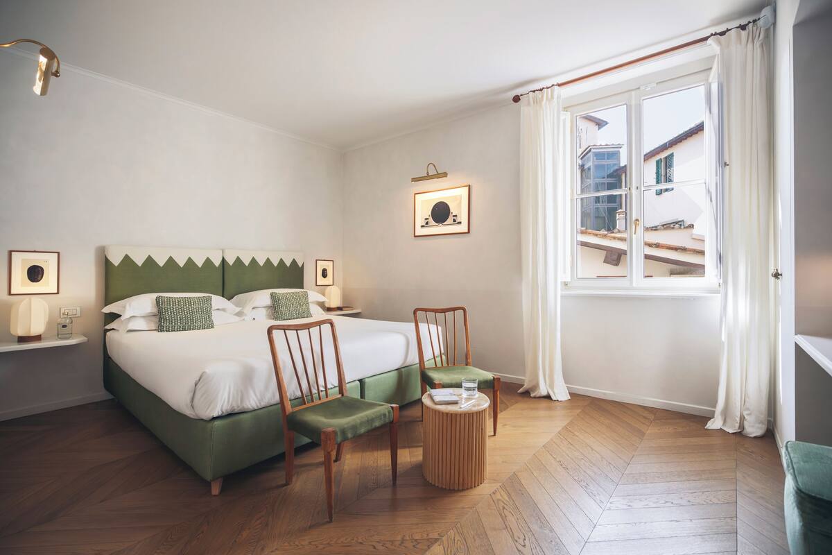 A hotel room with a bed, two chairs, a small table, a window with curtains, and a wall-mounted picture.