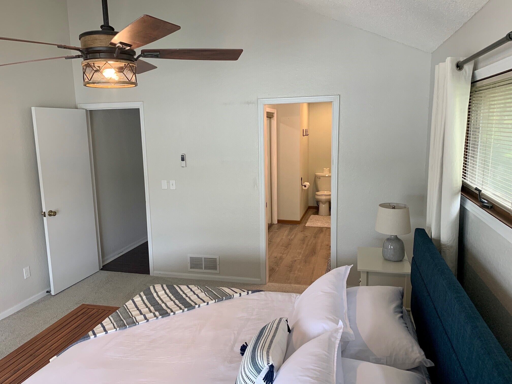 A bedroom with a bed, a ceiling fan, a nightstand with a lamp, and a view of the bathroom.