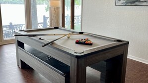 Une table de billard avec un cadre en bois et une queue de billard, dans une pièce dotée d’un plancher en bois et de grandes fenêtres.