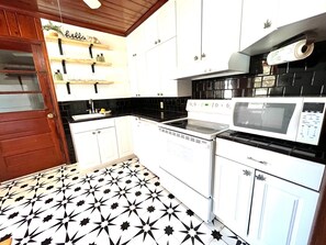 Compact kitchen with a stove, microwave, and sink, featuring a shelf with various items.