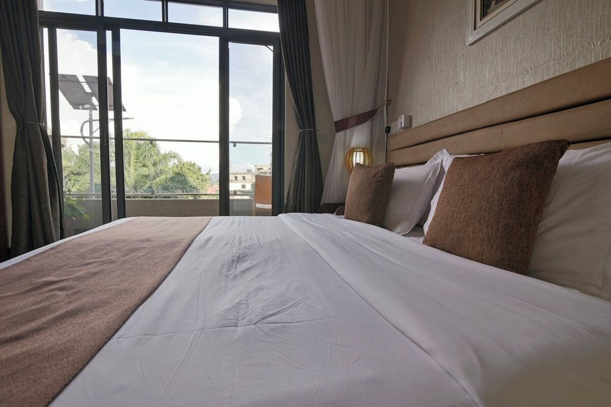 A hotel room with a large bed, a view of the city through the window, and a nightstand with a lamp.