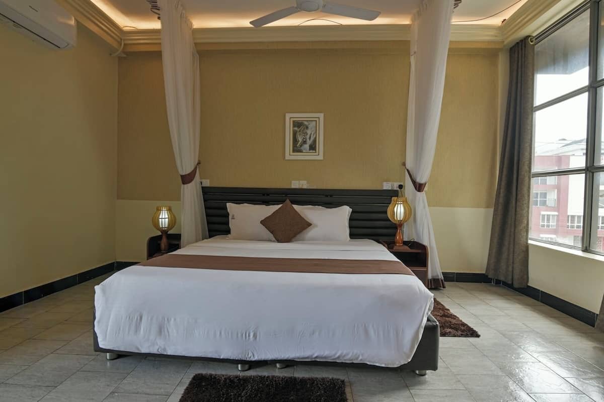 A large bed with white and brown bedding, two bedside lamps, a ceiling fan, and a large window with curtains.