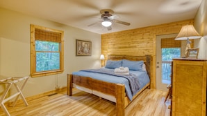 A bedroom with a wooden bed, a ceiling fan, a window with blinds, and a door with a view of trees.