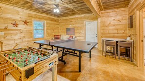 A wooden-floored room with a foosball table, ping pong table, and two bar stools.