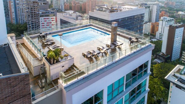 Una piscina en la azotea con sillas de descanso y barandilla de vidrio, rodeada de edificios altos.