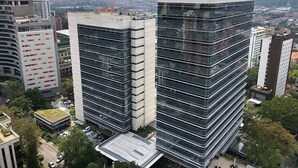Un moderno edificio de gran altura con fachada de vidrio, rodeado de otros edificios y un paisaje montañoso.
