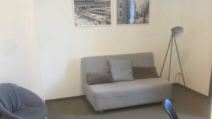 Une petite pièce au mobilier minimaliste, avec un canapé gris, un fauteuil gris et une chaise noire. Deux tableaux encadrés sont accrochés au mur.