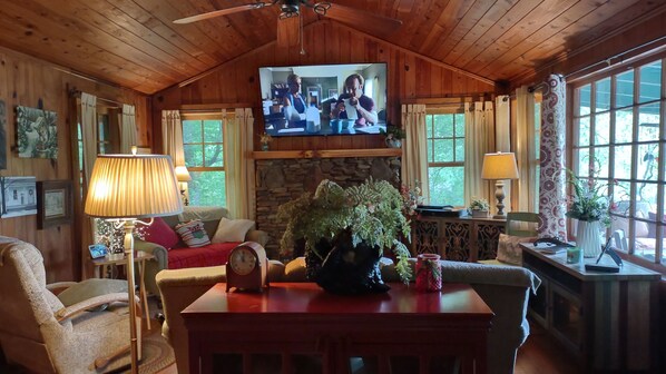 L’intérieur chaleureux d’un chalet, avec un téléviseur fixé au mur, une cheminée en pierre, des meubles en bois et de grandes fenêtres.