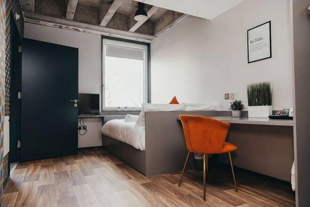 A compact bedroom with a bed, a desk, a chair, a window, and a door.
