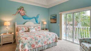 A bedroom with a mermaid mural, a bed with floral bedding, two bedside lamps, and a view through a sliding glass door. - Mermaid Beach House (St. Augustine)