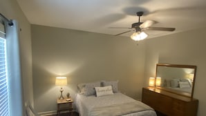 Une chambre à coucher comprenant un lit, une table de chevet, une commode, un miroir, un ventilateur de plafond et une fenêtre avec des stores.