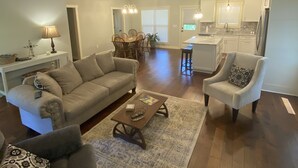 Un salon spacieux avec un canapé, des fauteuils et une table basse. Au fond, on aperçoit un coin repas avec une table et des chaises.