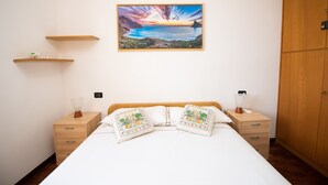 A neatly arranged bedroom with a bed, bedside tables, a nightstand, a wall-mounted shelf, and a framed picture of a landscape. - Casa Marisa (Toscolano Maderno)