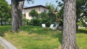 A grassy area with two white lounge chairs, a building with a red-tiled roof, and trees. - Casa Marisa (Toscolano Maderno)