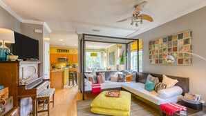 A living room with a sofa, ottoman, and a piano. There is a kitchen area with a microwave and a sink. - Bay Bridge Beach Boho (St. Augustine)