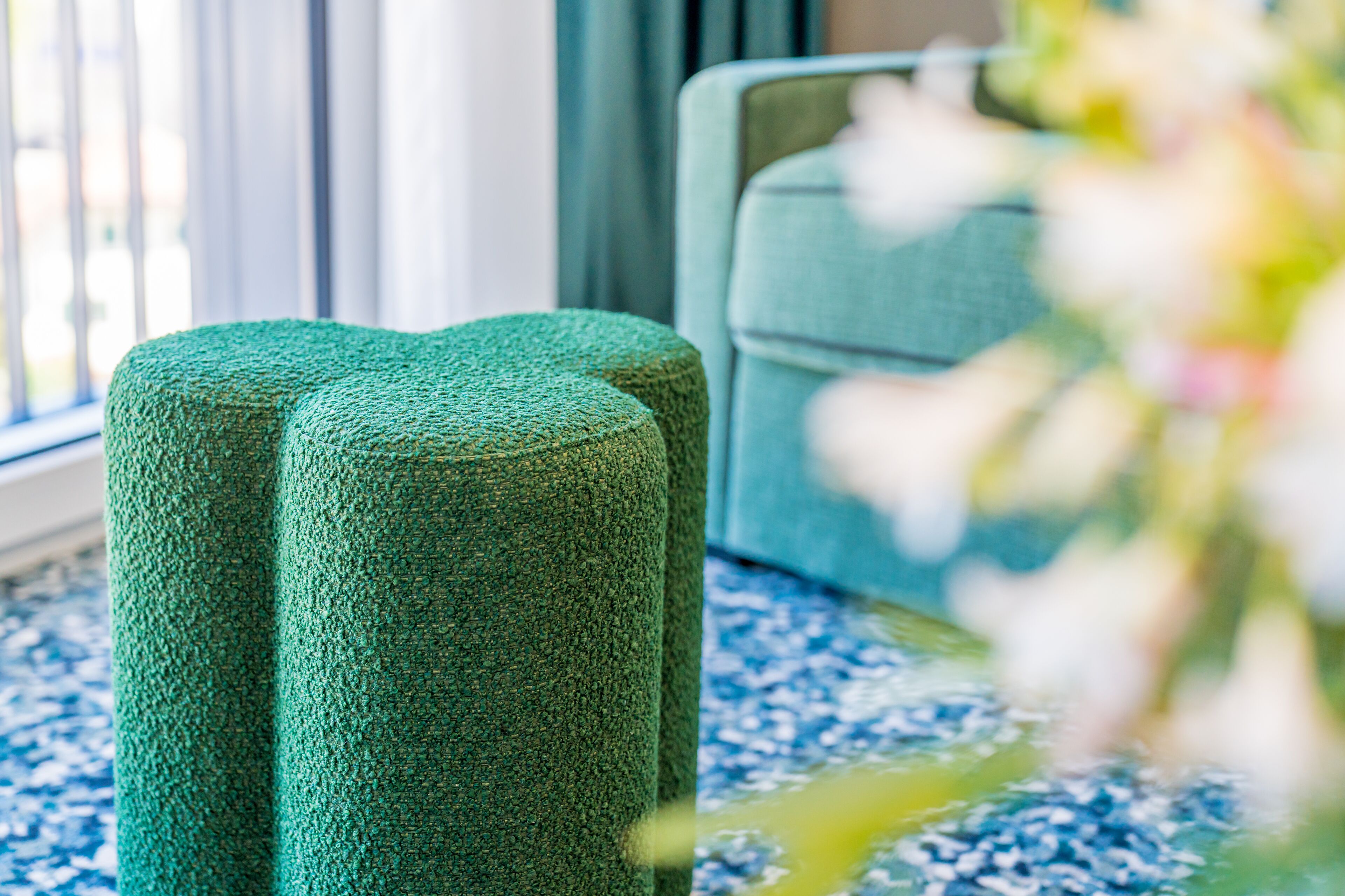 A room with a blue patterned carpet, a green textured ottoman, and a light blue armchair.