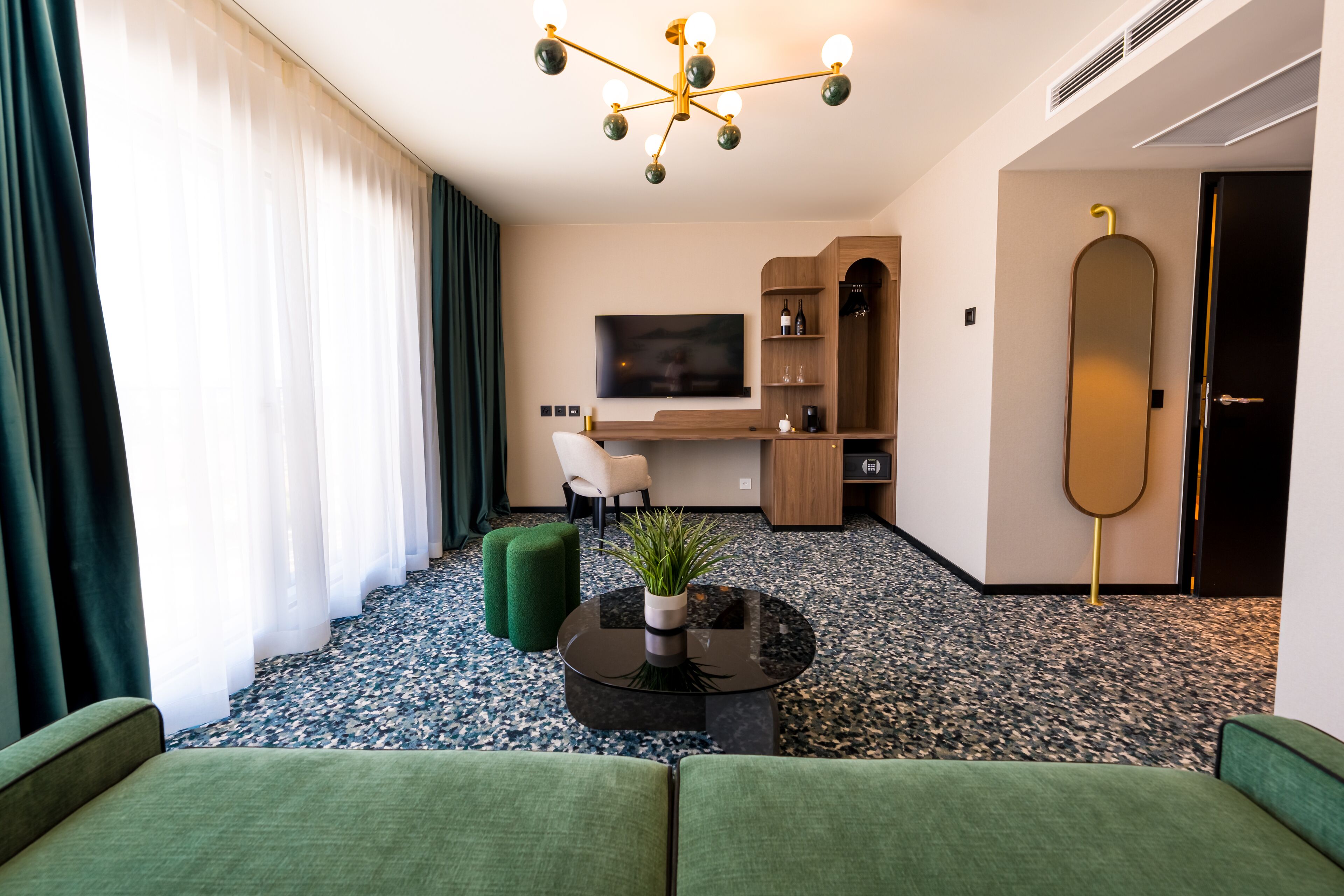 Ein modernes Hotelzimmer mit einem grünen Sofa, einem schwarzen Couchtisch mit Pflanze, einem Fernseher, einem Holztisch und einem großen Spiegel.
